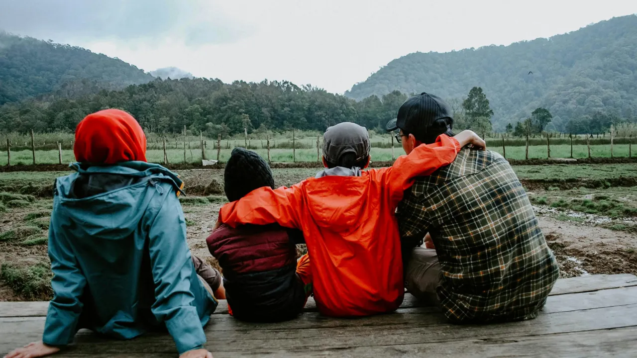 A family sitting close on a wooden deck, arms around each other, looking out at misty green hills
